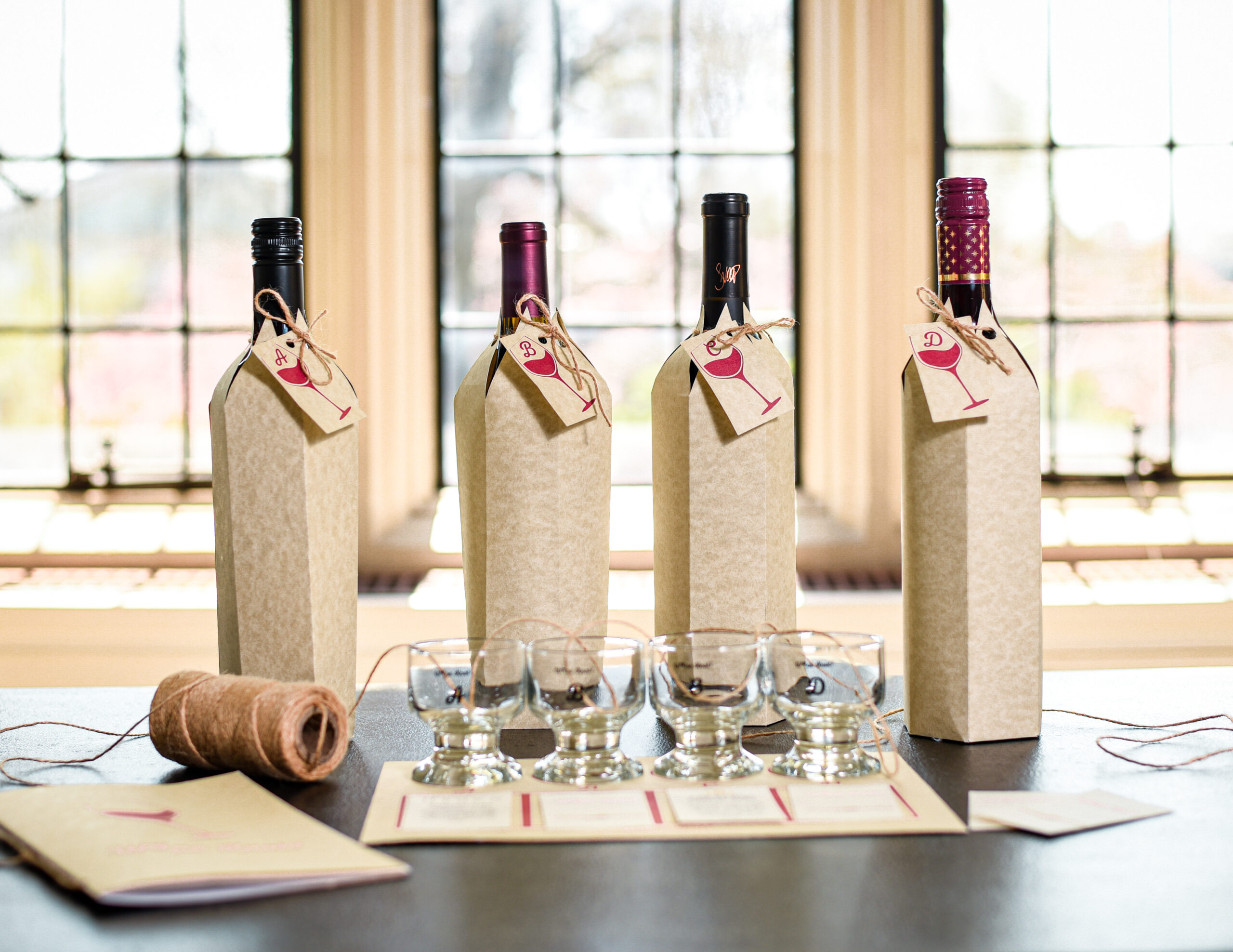 Four bottles of wine covered in a cardboard sleeve to conceal the label sit on a wooden table in a light filled room. Four small tasting glasses sit in front of the bottles of wine.