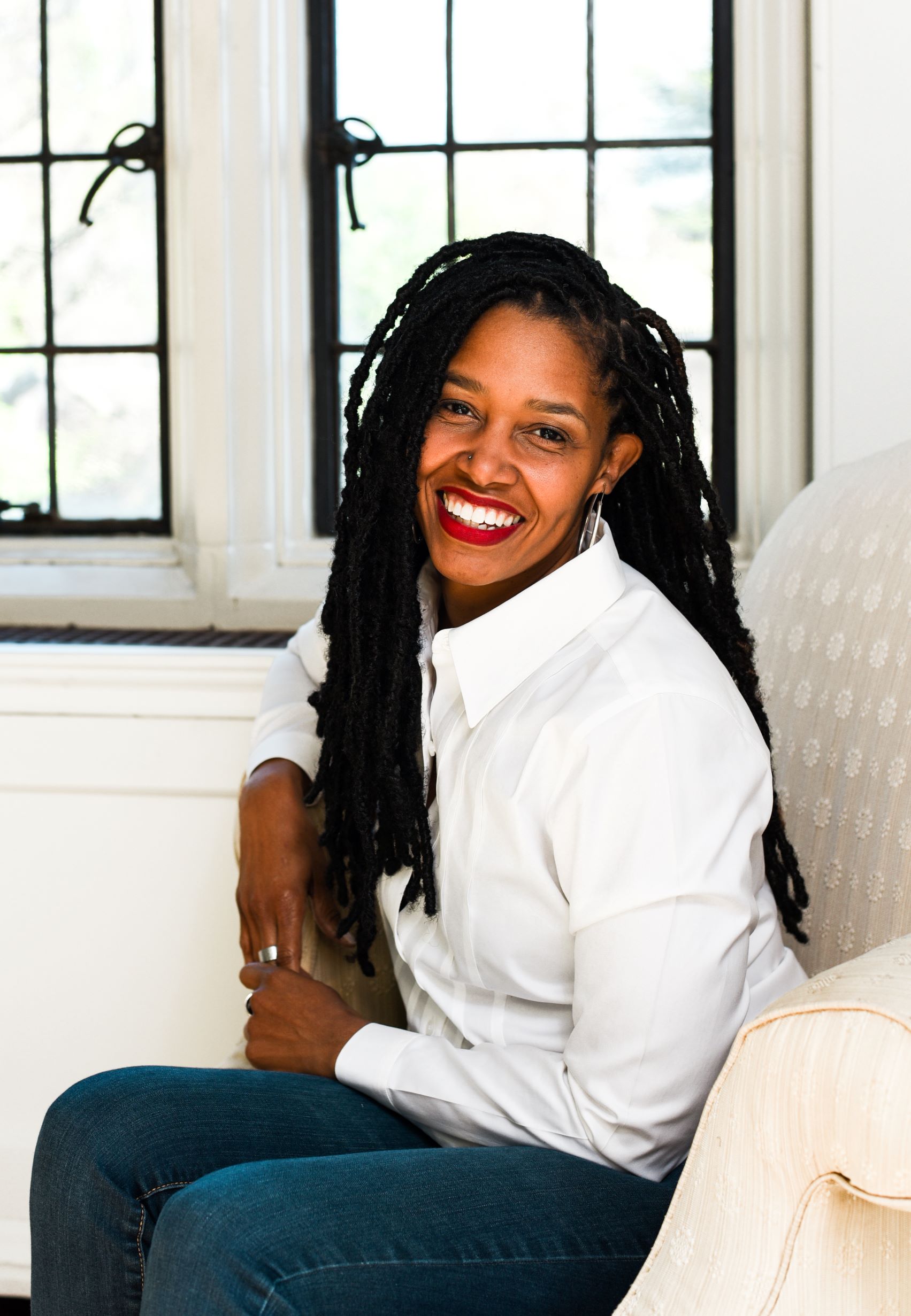 Pamela Weaver, a Black woman, is sitting on a antique white couch in a white oxford shirt, jeans, red lipstick, and a relaxed smile